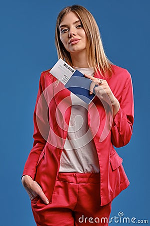 Blonde lady in white blouse and red pantsuit. She smiling, holding passport and ticket while posing on blue studio Stock Photo