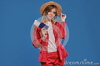 Blonde model in straw hat, white blouse, red pantsuit. She is holding passport and ticket, posing on blue background Stock Photo