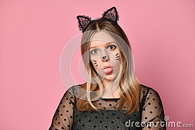 Blonde teenager in black dress, headband like cat ears, face painting. She showing her tongue, posing on pink background. Close up Stock Photo