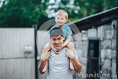 Blonde three years old boy sitting on father`s shoulders. Kazakh father and caucasian mother. Happy father with black hair play Stock Photo