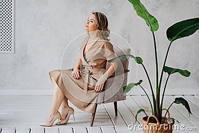 A blonde woman in a beige dress poses on a gray chair in a photo studio. Stock Photo