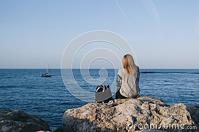 A blonde woman on her back sitting on a rock by the sea watching a sailboat sail a sunny day Stock Photo