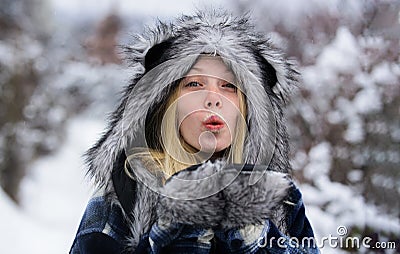 Blonde woman in plaid coat, fur hat and mittens in winter park sending air kiss. Portrait of beautiful girl in warm Stock Photo