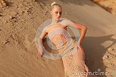 Gorgeous blonde woman in gorgeous golden dress posing in the desert, lying on the sand at sunset Stock Photo
