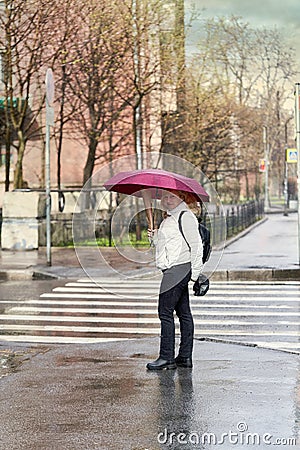 Blonde woman in a white jacket and black jeans covers herself with red umbrella Stock Photo