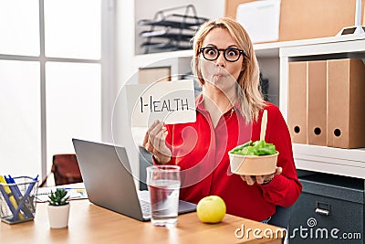Blonde woman working at the office eating healthy food making fish face with mouth and squinting eyes, crazy and comical Stock Photo