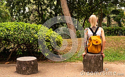 A blonde with a yellow backpack sits in a green park. Wears a red bandage, arms with tattoos. Asia travel Stock Photo