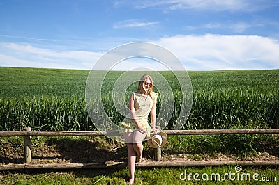 Blonde, young, pretty farm woman, sitting on a wooden fence. In the background a green wheat field. The woman is wearing a short Stock Photo