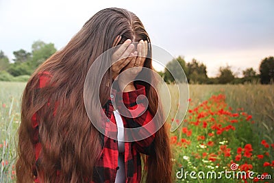 Blooming red poppies in summer field, girl with long hair stands, covering her face with her hands and crying, concept of Stock Photo