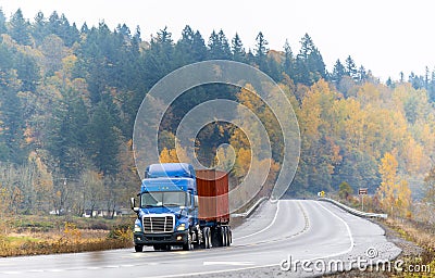 Blue big rig semi truck with flat bed semi trailer transporting container running on winding autumn road with raining weather Stock Photo