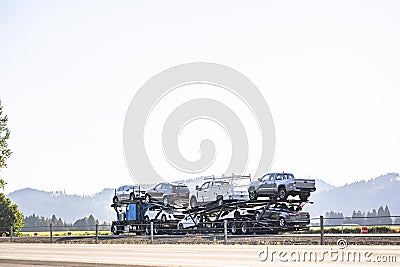 Blue classic old style big rig car hauler semi truck tractor transporting cars and crossovers on hydraulic semi trailer driving on Stock Photo