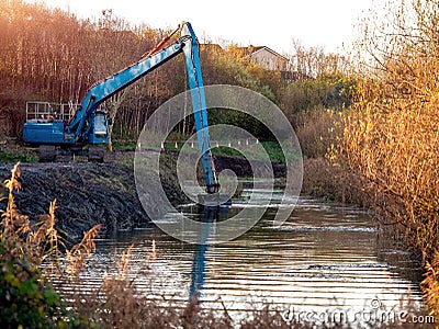 Blue excavator cleaning river in a park, Warm morning light Stock Photo