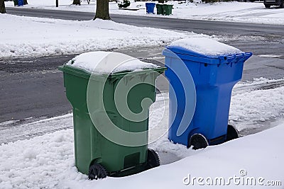 A blue and a green trash can covered with snow by the side of a snow covered, freshly plowed street waiting for garbage pickup Stock Photo
