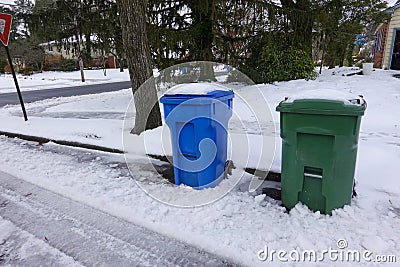 A blue and a green trash can covered with snow by the side of a snow covered, freshly plowed street waiting for garbage pickup Stock Photo