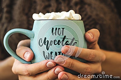 Blue mug with coffee, hot chocolate or cocoa with marshmallow in female hands. Inscription on cup Hot chocolate weather Stock Photo