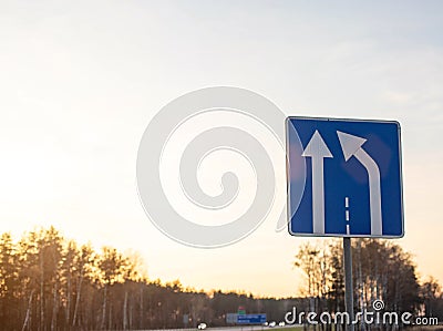 Blue road sign on sky background with sunny sunset narrowing the road to the right. Concept of warning signs about lane change, Stock Photo