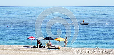 Blue sea with boats and beach with four colorful umbrellas and sitting people Editorial Stock Photo