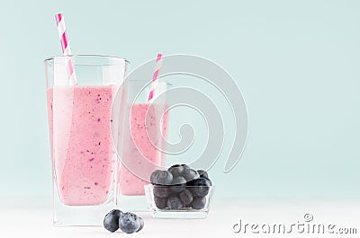 Blueberry milkshakes in transparent glass with blueberries in bowl, striped straw on white wood table and mint color wall. Stock Photo