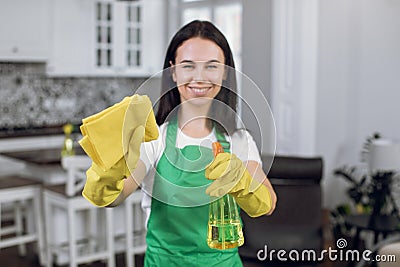 Blurred view of pretty smiling young female cleaner in gloves and apron, spraying cleaner detergent to camera and making Stock Photo