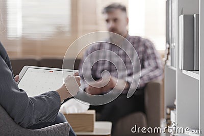 Blurred view of a single man sitting on a sofa during a psychotherapy session with a side view of the adviser holding a tablet in Stock Photo