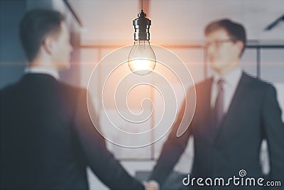 Blurry businessmen shaking hands together on abstract office interior background with glowing light bulb. Work ideas and Stock Photo