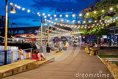 Blurry people hanging out at waterfront boardwalk string lights, beach chairs, patio restaurant dining tables sunset blue hour Editorial Stock Photo