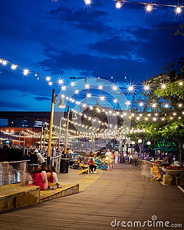 Blurry people hanging out at waterfront boardwalk string lights, beach chairs, patio restaurant dining tables sunset blue hour Editorial Stock Photo