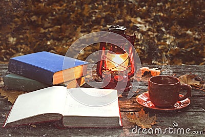 The book, lamp and a cup of hot coffee on the old wooden table in a forest. Fallen yellow maple leaves. Stock Photo