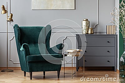 Books on stylish golden small table next to emerald green velvet wing back chair in grey living room interior Stock Photo