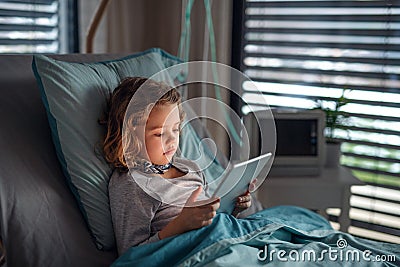 Bored small girl in bed in hospital, using tablet to pass time. Stock Photo