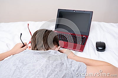 Bored and tired boy lying on a bed with a laptop after doing his homework, distant remote education during quarantine Stock Photo