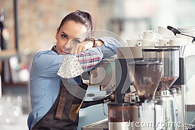 Bored waitress leans against the coffee machine with no job to do, holding face mask in her hand Stock Photo