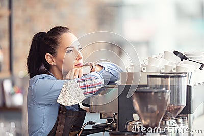 Bored waitress leans against the coffee machine with no job to do, holding face mask in her hand Stock Photo