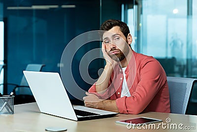 Boring office work. A young man sits in the office at a table with a laptop, leans on his arm and is bored, tired, looks Stock Photo