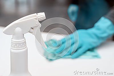 Bottle with disinfection spray in the foreground, blurry person with blue gloves in the back cleaning the white surface, to Stock Photo