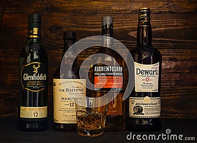 Bottles of 12 year old Balvenie, Auchentoshan, Glenfiddich, Dewars whiskey and a glass with ice on a dark wooden Editorial Stock Photo