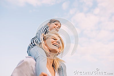 Bottom view of cute little daughter on a piggy back ride with her smiling mother on sky background. Loving woman and her little Stock Photo