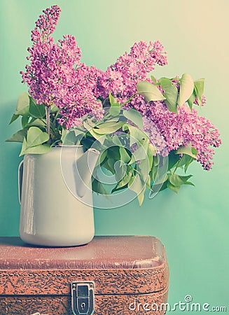 Bouquet of lilac in an old enameled teapot on vintage suitcase on blue background. Retro style still life. Stock Photo