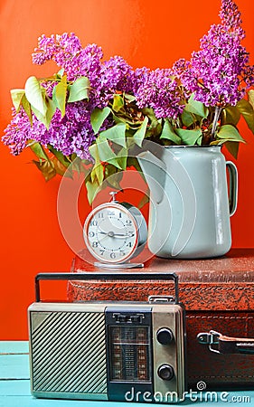 Bouquet of lilacs in enameled kettle on antique suitcase, vintage radio, alarm clock on yellow background. Retro style still life. Stock Photo