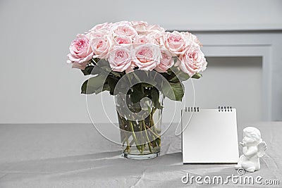 A bouquet of pink double roses in a glass transparent vase on a table covered with a gray tablecloth. Notebook. And a statuette of Stock Photo