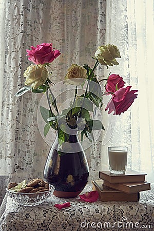 A bouquet of roses in a ceramic vase . Next to it are books , a glass of milk, and a bowl of cookies Stock Photo