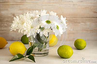 Bouquet of white chrysanthemums in glasswith whole lemons and limes. Abstract floral and fruits rustic background. Stock Photo