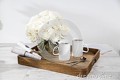 Bouquet of white roses in a round glass vase on a tray with two cups of tea and spoons in the interior Stock Photo