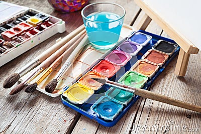 Box of watercolor paints, art brushes, glass of water and easel Stock Photo