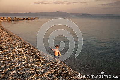 Boy on the beach. concept of loneliness. Cute child waving hand at seashore in evening. Boy sits in sea water, rear view Stock Photo