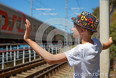 A boy in a beautiful colored hat with a smile waves to a train passing by on the railway, seeing off or meeting passengers Stock Photo