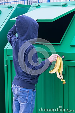 A boy in a blue hooded jumper throws a banana skin into a green dumpster Stock Photo