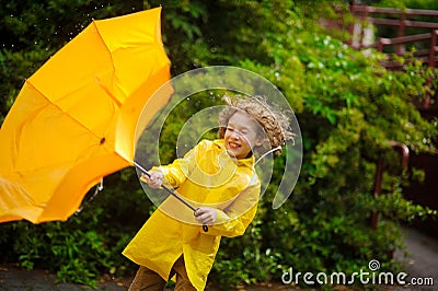 The boy in a bright yellow raincoat with effort holds an umbrella from wind. Stock Photo