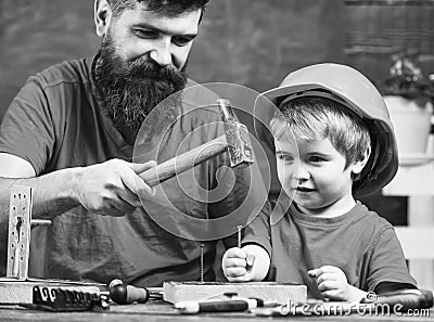 Boy, child busy in protective helmet learning to use hammer with dad. Father with beard teaching little son to use tools Stock Photo