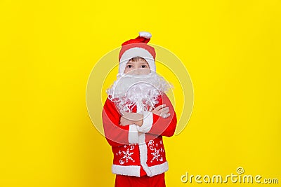 A boy in a .d santaclaus suit and an artificial beard stands on a yellow background and crosses his arms on his chest Stock Photo
